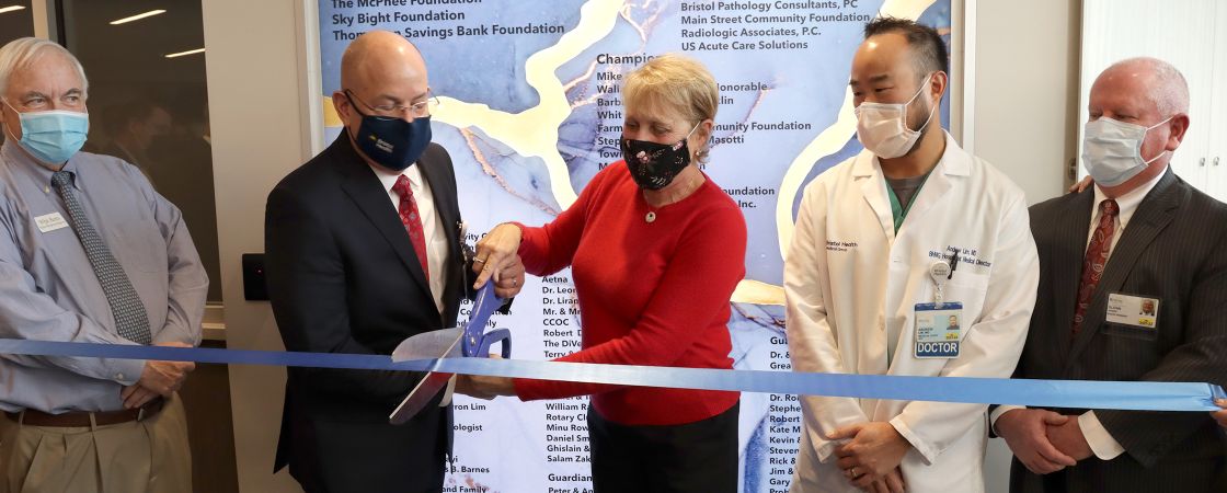 A woman in a red shirt and man in a suite cutting a blue ribbon with scissors while standing next to a doctor.