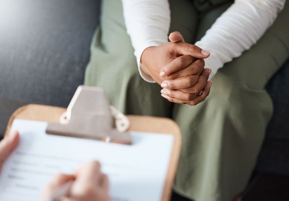 Close-up of a counselor taking notes on a clipboard while the patient sits across on a couch with hands folded